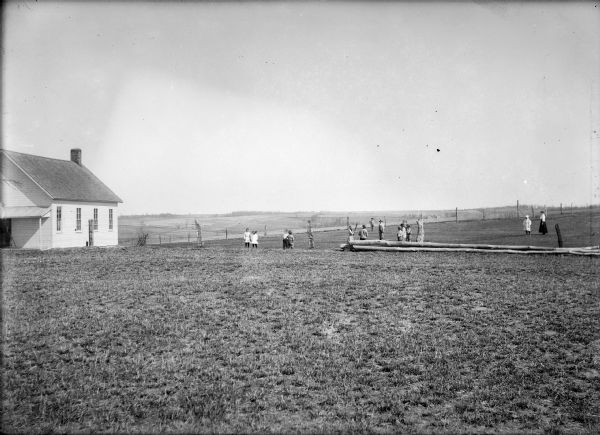 View across lawn of a clapboard one-room schoolhouse with students and a teacher playing baseball on the right in the middle distance.
