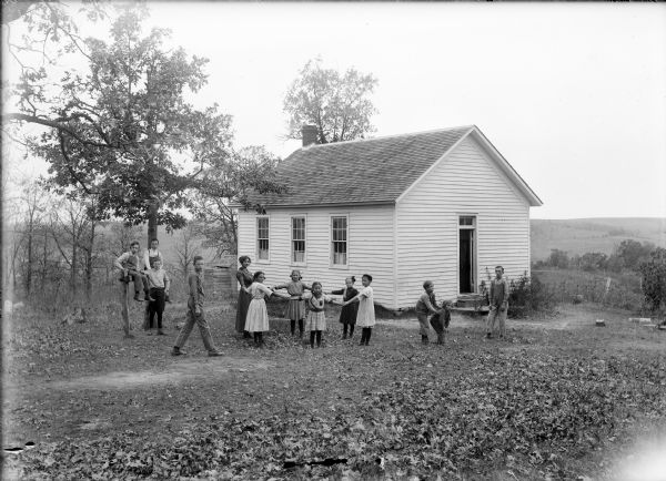 View across yard of a clapboard one-room schoolhouse with a teacher and children playing outside.