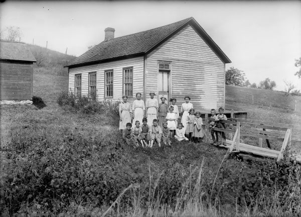 Students posed around a wood bridge outside a clapboard one-room schoolhouse. Small building on left may be an outhouse. In the background is a hill with a fence.
