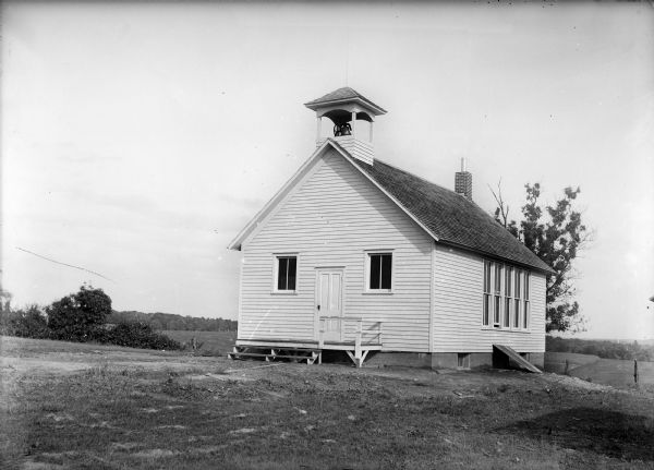 One-room clapboard school building with bell tower. Field and trees are in the background.