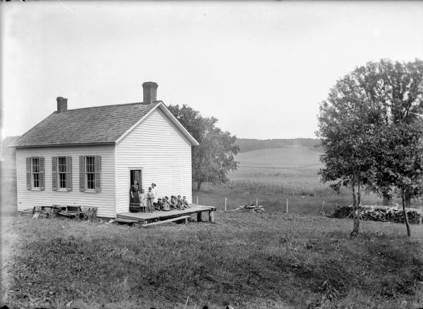 View down hill of a teacher standing in an open doorway, with students standing and sitting on the wooden landing of a clapboard one-room school building. There is a large wood pile along the fence on the right, and fields and trees are in the background.