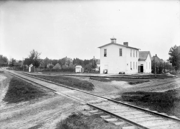 View across railroad tracks of a two-story clapboard schoolhouse with bell tower. Groups of children pose outside and another group of children look out one of the second-story windows. An outhouse is visible behind the school.