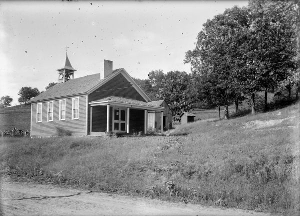View from road of a clapboard one-room school building with white trimming and a cream-colored chimney. There is a bell tower with a weather vane on top. An outhouse is in the background on a hill.