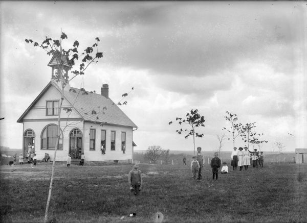 A teacher, students, and a dog are carefully arranged on and around a two-story clapboard school building and school grounds. Young trees are planted on the lawn. There is a bell tower on the roof, two arched doorway entrances, and a large arched window on the school building.