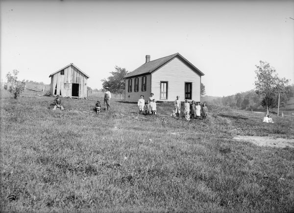 Students with their teacher pose on the grounds in front of a clapboard one-room schoolhouse and shed.