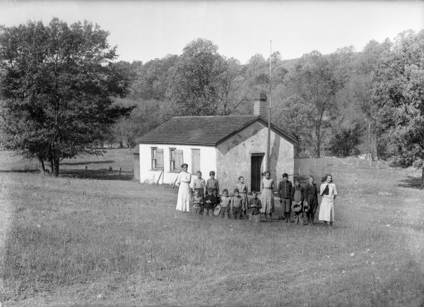 View down slope of a teacher and students posing in front of a one-room school building with a plaster exterior. Behind the school are a fence, trees, and a hill.