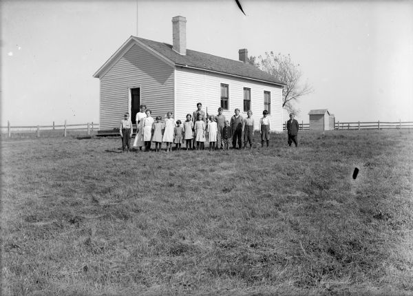 A group of students pose in front of one-room schoolhouse. Surrounding the grounds is a wood fence, and there is an outhouse on the right.