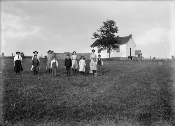 Teacher and students pose outside on the grounds near a one-room schoolhouse.