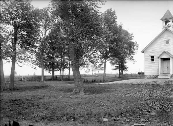 View of school grounds with school building with small bell tower on the right. In the background is a fence, and a man driving a wagon.