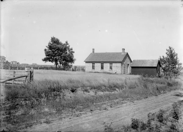 View from road of a stone one-room school house and shed. In the background is a field and farm buildings.
