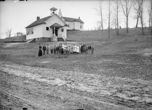 View from dirt road of teacher and students posed outside a clapboard schoolhouse with a front door gable roof and a bell tower. On the left side of the school building is an exposed stone foundation with a bulkhead. In the background on a hill is a church.