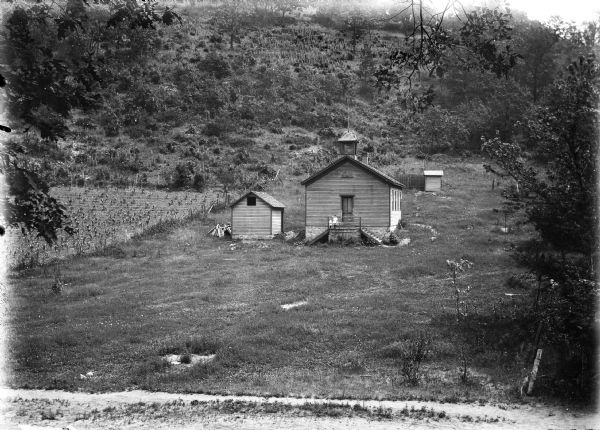 Clapboard one-room schoolhouse with bell tower. There is also a shed, outhouse, and a small field with a fence on the school grounds. Two children pose on landing in front of the door. A hill rises steeply in the background.