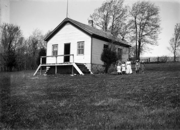 View up slope of unidentified clapboard one-room schoolhouse with teacher and students posed outside. In the background is a fence and field.