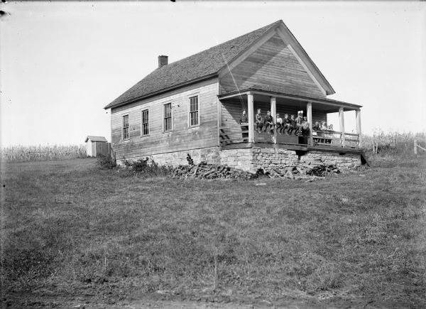 View up hill of clapboard school building with stone foundation. The teacher and students are posed along the railing of the porch. In the background is a fence and outhouse.