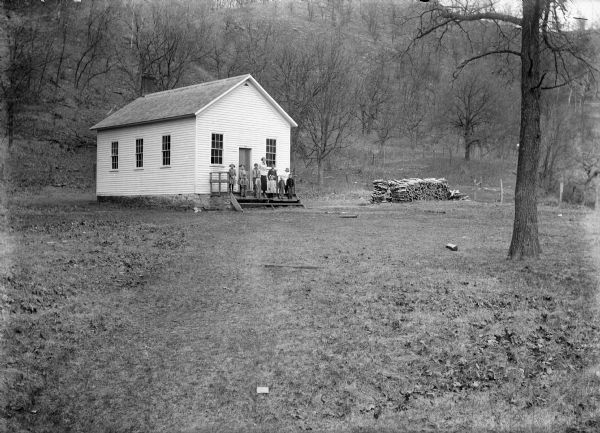 A teacher and students are posing on the landing of a clapboard one-room schoolhouse. On the right is a stack of firewood, and a hill rises steeply in the background.
