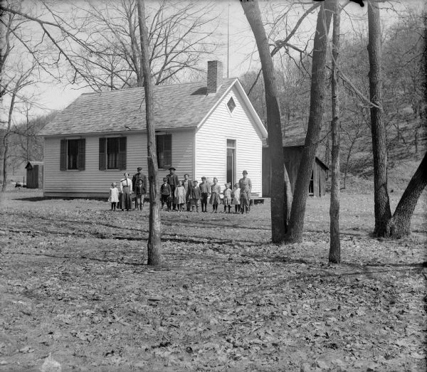 View through trees of a group of children and adults standing in front of a clapboard one-room schoolhouse. an outhouse is visible behind the shcool.