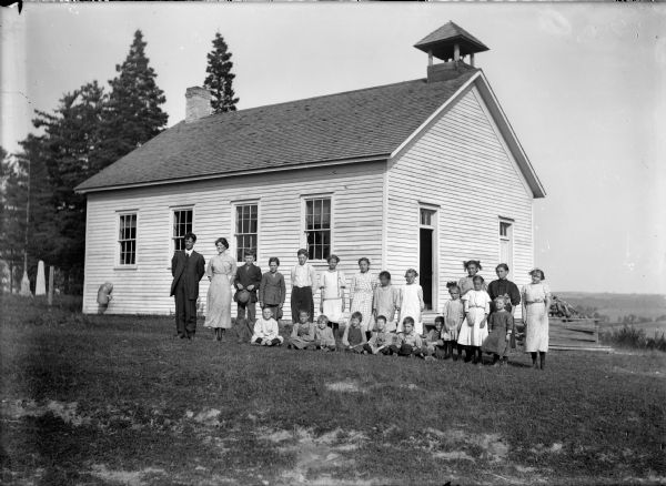 A teacher and students pose outside a clapboard one-room schoolhouse with small bell tower.