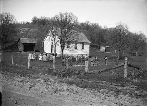 View from road of students and teachers posing in front of a clapboard  one-room schoolhouse. Some of the boys wear baseball gloves. A hill rises in the background. An outhouse is visible behind the school.