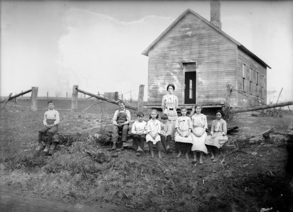 Teacher and students pose on logs in front of a clapboard one-room schoolhouse.