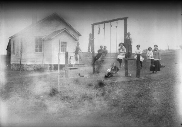 Teacher and students pose on and around playground structure with swings outside a clapboard one-room schoolhouse.