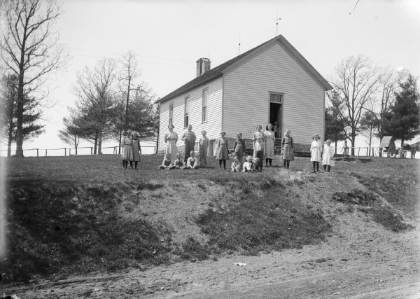 View from road up hill of teacher and students posing in front of a clapboard and one-room schoolhouse. A horse and buggy are on the hill on the right.