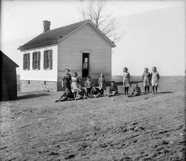 Teacher and students posing outdoors in front of a one-room clapboard schoolhouse with a stone foundation. A small building is on the left.