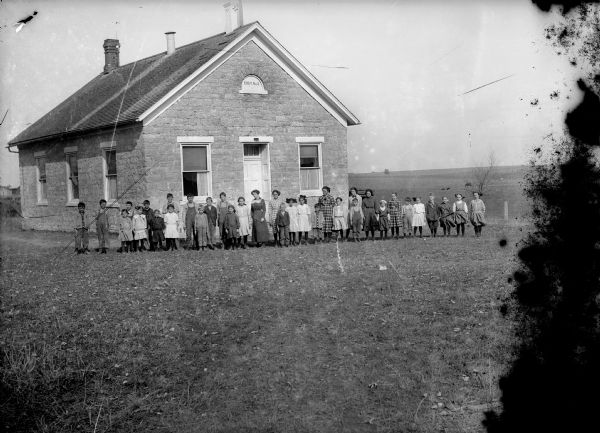 A teacher and a large group of students are posing in front of a stone one-room schoolhouse. A sign above the entrance says: "DIST. No2."