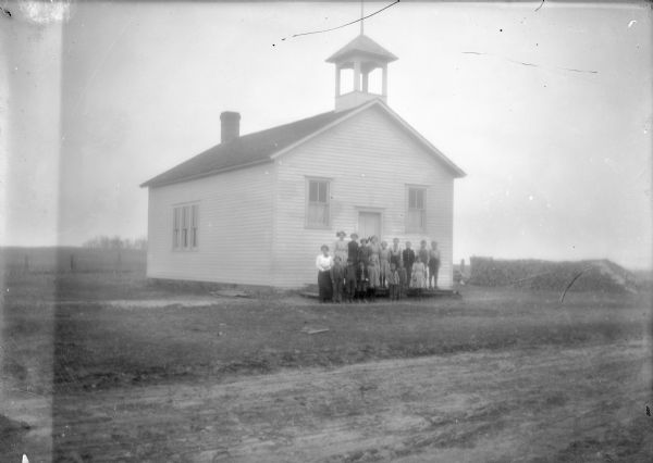 View from road of a teacher and students posing on front landing of a clapboard one-room schoolhouse with bell tower.