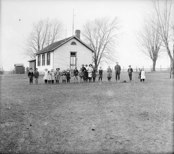 Students pose outdoors in front of a clapboard one-room schoolhouse. In the background are an outhouse, fence, and trees.
