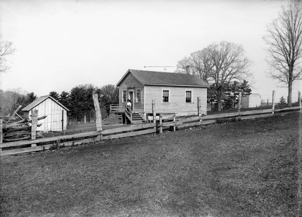 View over fence of a teacher wearing a large, dark hat posing on the front landing of a clapboard one-room schoolhouse.