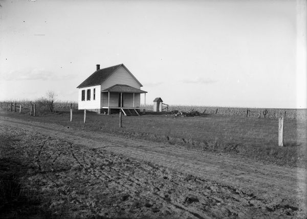 View from dirt road of a clapboard one-room schoolhouse with porch. There is an outhouse behind it near a field. The school was closed as children were given $25 each to attend school in another district.