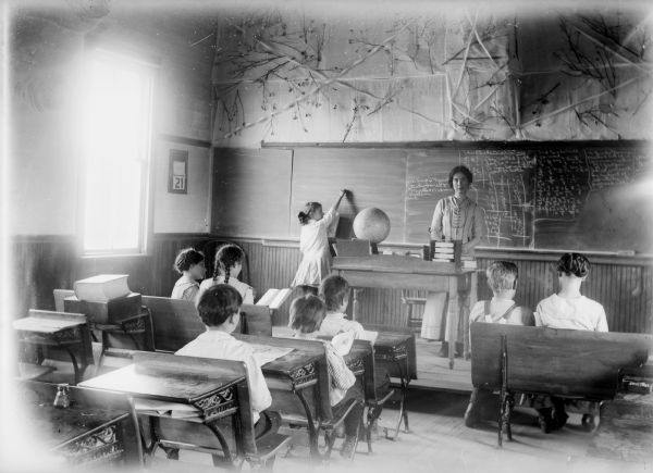 View from back of classroom of teacher and students during a class. There is a student standing at the chalkboard, and the teacher is standing behind her desk. The wall above the chalkboard is decorated with tree or shrub branches.