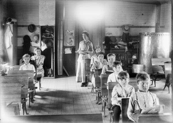 View from front of room of teacher and students in classroom of one room schoolhouse. There is a large stove on the right, and the teacher stands behind the students posing at their desks.