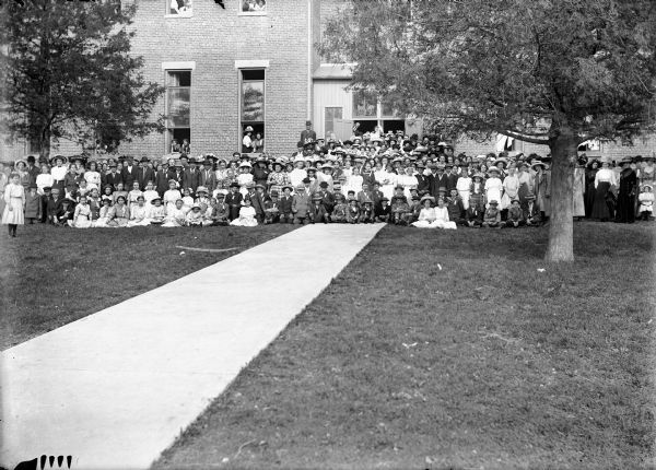 Large group of children and adults pose on the lawn in front of brick building. Other people look out of open windows.