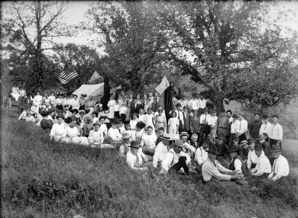 A large group of children and adults pose in a field around trees. There is a tent in the background with flags, and two women sit in a carriage on the left. Edward Parker, teacher.