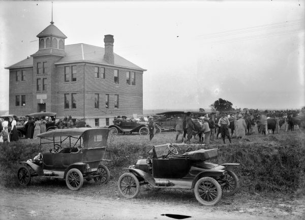 View from the road of fairgoers on front lawn of Union High School. Two automobiles are parked in the foreground.