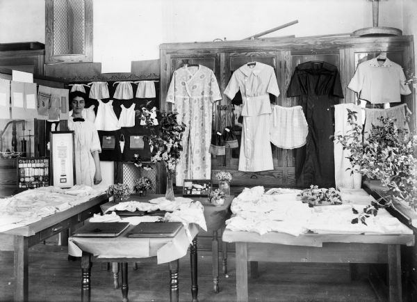Woman standing at sewing exhibit that includes dresses, children's clothes, aprons, and hats. Mathilda Monteith was the instructor of the sewing class.