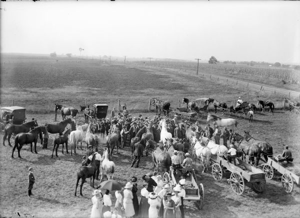 Elevated view of crowd around horses during livestock judging. In the background are fields, a road, power lines, and in the far distance along the horizon a windmill.