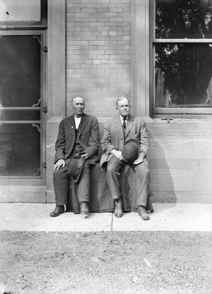 Two men sit outside on bench on a sidewalk in front of a building near a door.