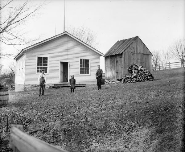 Two unidentified men and a young girl posing outside what is possibly a one-room schoolhouse.