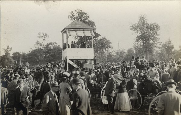 Large crowd of people around a viewing platform at what appears to be a horse race or possibly a sporting event.