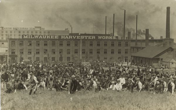 Exterior view of the Milwaukee Harvester Works. View from hill towards a large crowd of workers, mainly men and boys, posing in front of the factory in an open field.