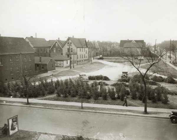 Elevated view of entrance to National Park on a city block. A road passes by a grove of evergreen trees lined up along the sidewalk, while two men work near stacks of other evergreens.