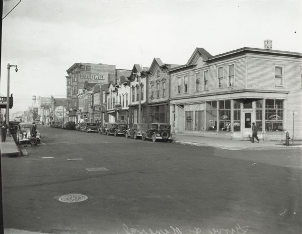 Corner of Grove and Mineral Streets with cars parked along the road. Storefronts line the streets. The Goodwill store, a used car dealership and a water tower are in the background. A man with a broom and a wheelbarrow stands in the street near the corner.