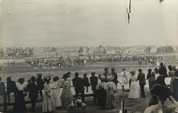 A group of spectators including men, women, and children standing at a railing watching some type of sporting event in a field where a large crowd is gathered. There are houses in the background. In the foreground women are wearing dresses and large hats and nearby is a baby in a carriage.