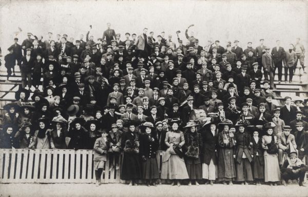 Group portrait of a crowd of men, women, and children standing on bleachers for a sporting event. In the lower right-hand side there are several boys who have musical instruments, including drums and trumpets.