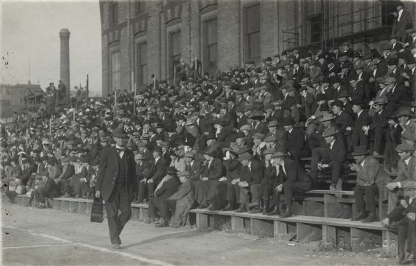 A crowd of mostly men sitting on bleachers in front of a brick building, all watching some type of sporting event. One man is walking in front of the bleachers and is carrying a case of some kind in his right hand.