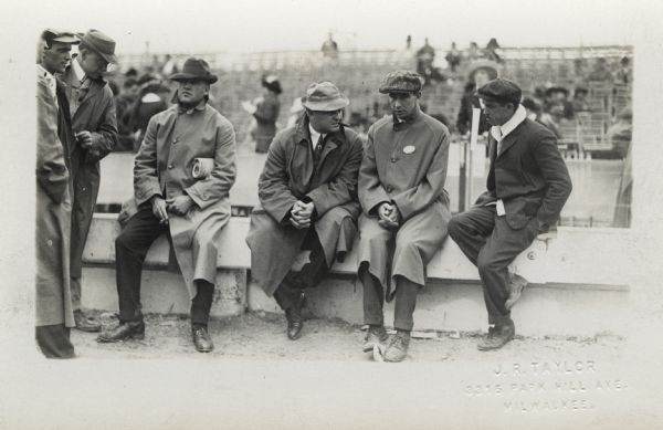 A group of six men sitting and standing near a barrier wall at a sporting event with bleachers in the background. They are all wearing jackets and hats, and three men are smoking.