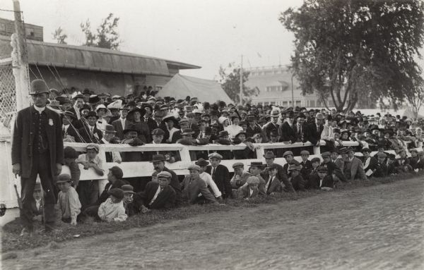 View from track towards a crowd of men, women, and children in front of and behind a fence at a sporting event. On the left, a man who is possibly a police officer, is standing and keeping control over the crowd.