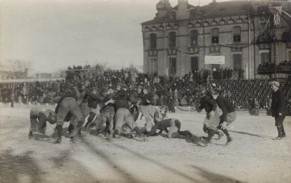 View of men on field during football game. Possibly taken in Milwaukee, Wisconsin. In the background a crowd is watching on stands in front of a large brick building.